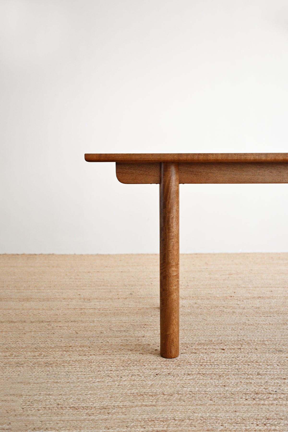 A close-up of a wooden table leg and edge on a light beige woven rug against a plain off-white wall.