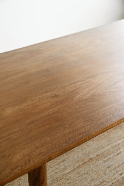 A close-up view of a warm medium-brown wooden table surface with visible wood grain, shown in a softly lit interior setting with a light neutral wall and woven rug.