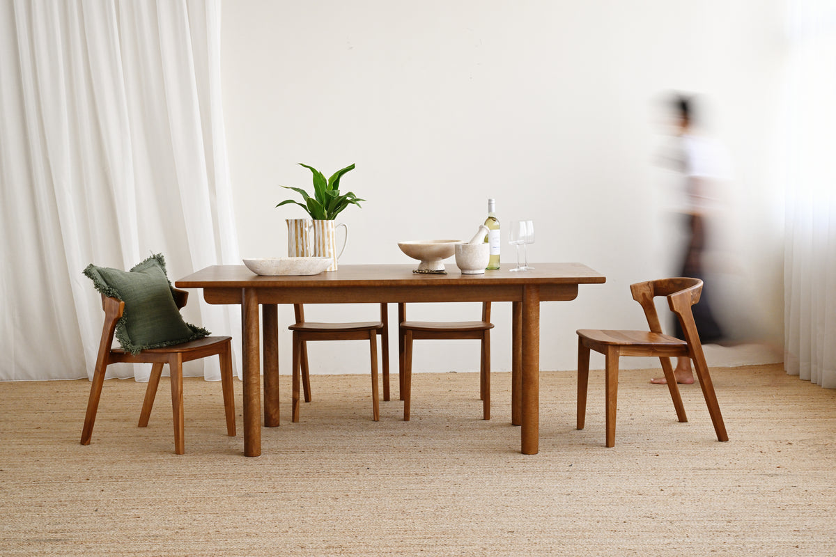 A wooden dining table with four matching wooden chairs in a bright, minimalist room with white curtains and a woven jute rug. A green pillow rests on one chair, and a blurred person is visible in the background near the window.