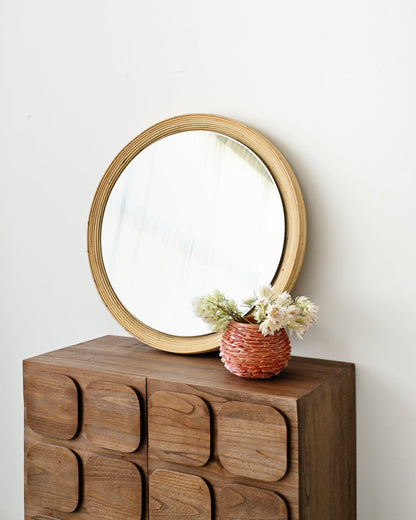 A round wooden-framed mirror leaning against a white wall on top of a dark brown wooden chest with a wavy front, styled with a small woven vase of white flowers.