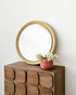 A round wooden-framed mirror leaning against a white wall on top of a dark brown wooden chest with a wavy front, styled with a small woven vase of white flowers.