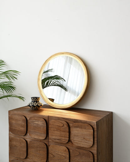 A round wooden-framed mirror with a gold-toned frame rests on top of a dark brown wooden cabinet with a unique raised geometric pattern, set against a plain white wall.