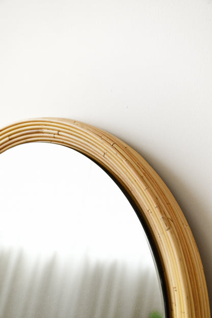 A close-up of a round mirror with a natural light wood or rattan frame against a plain white wall.