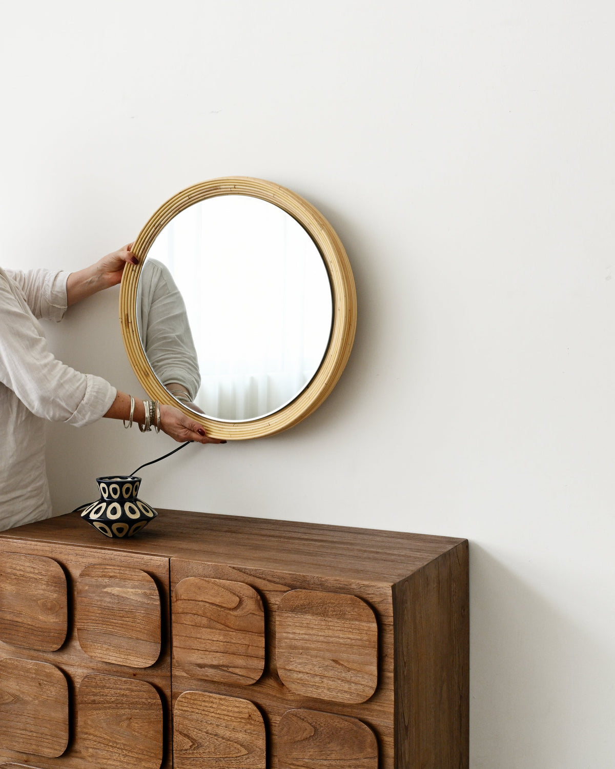 A woman in a white outfit hangs a round wooden-framed mirror on a white wall above a dark brown wooden chest with decorative raised drawer fronts.