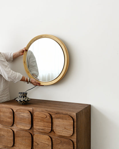 A woman in a white outfit hangs a round wooden-framed mirror on a white wall above a dark brown wooden chest with decorative raised drawer fronts.