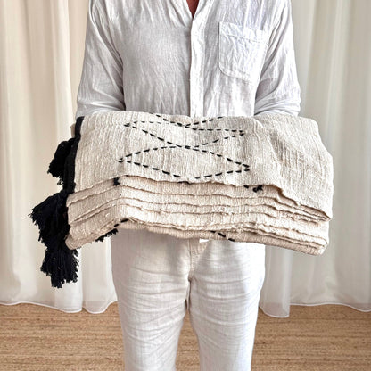 A person wearing a white linen shirt and white pants holds a folded cream-colored textured blanket with black embroidered geometric patterns and black tassels.