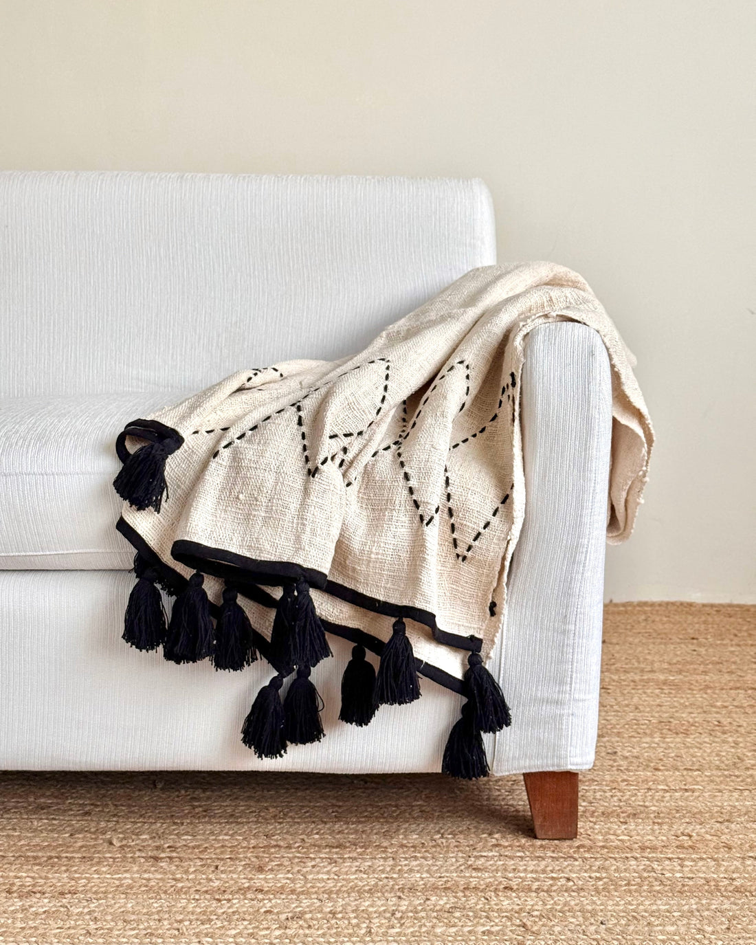 A cream-colored woven throw blanket with black tassels and embroidered geometric patterns, draped over the arm of a white sofa.