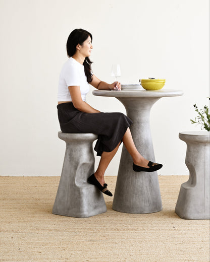 A woman sits on a gray concrete bar stool at a matching round concrete bar table, wearing a white t-shirt and a dark skirt. The setting is a minimalist room with a light beige wall and a textured beige floor.