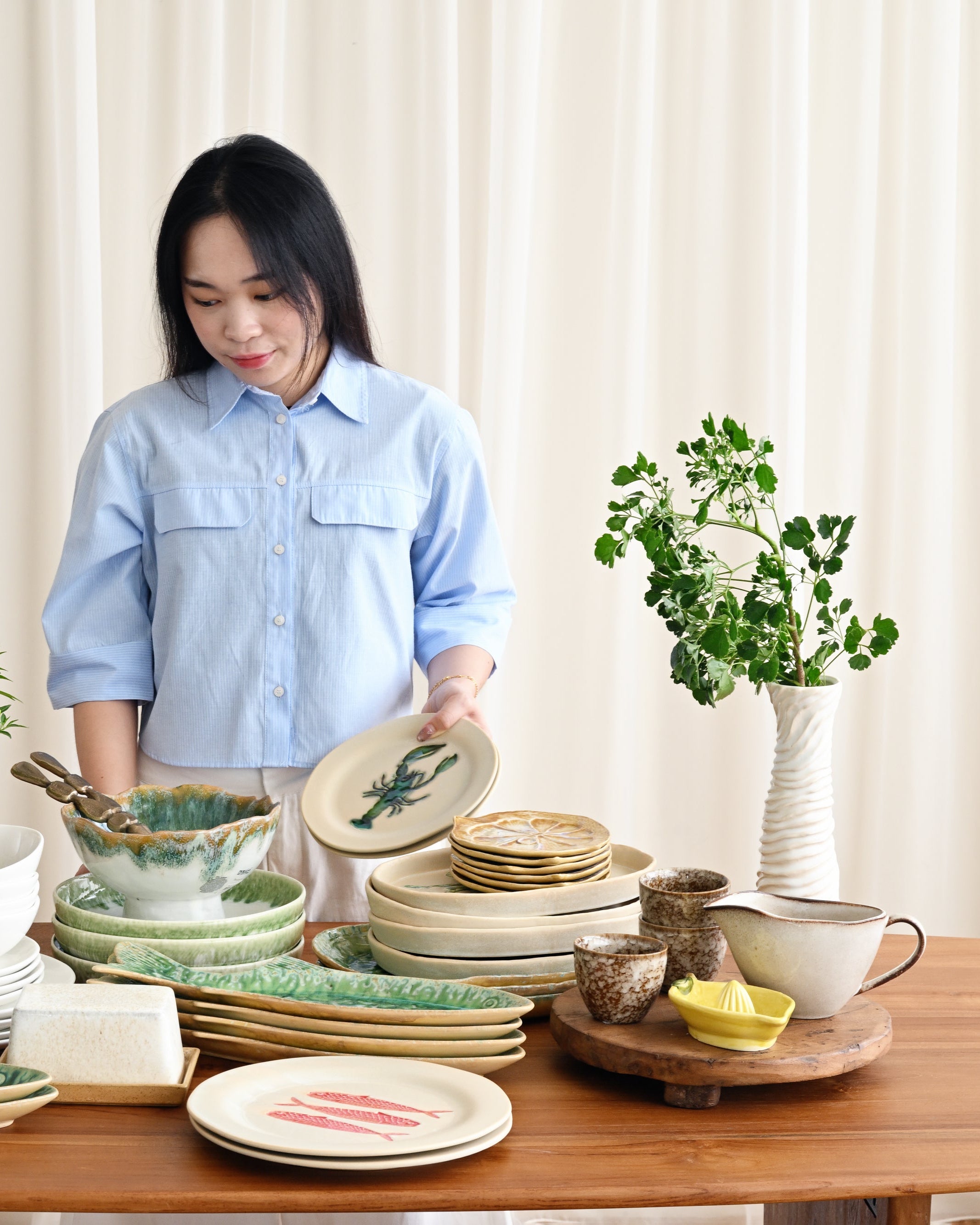 Woman arranging ceramic plates and bowls on a wooden table with plants in the background.