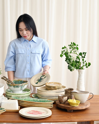 Woman arranging ceramic plates and bowls on a wooden table with plants in the background.