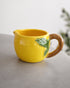 A yellow ceramic pitcher shaped like a lemon with a green leaf detail and a brown handle, set on a gray surface against a light gray background.