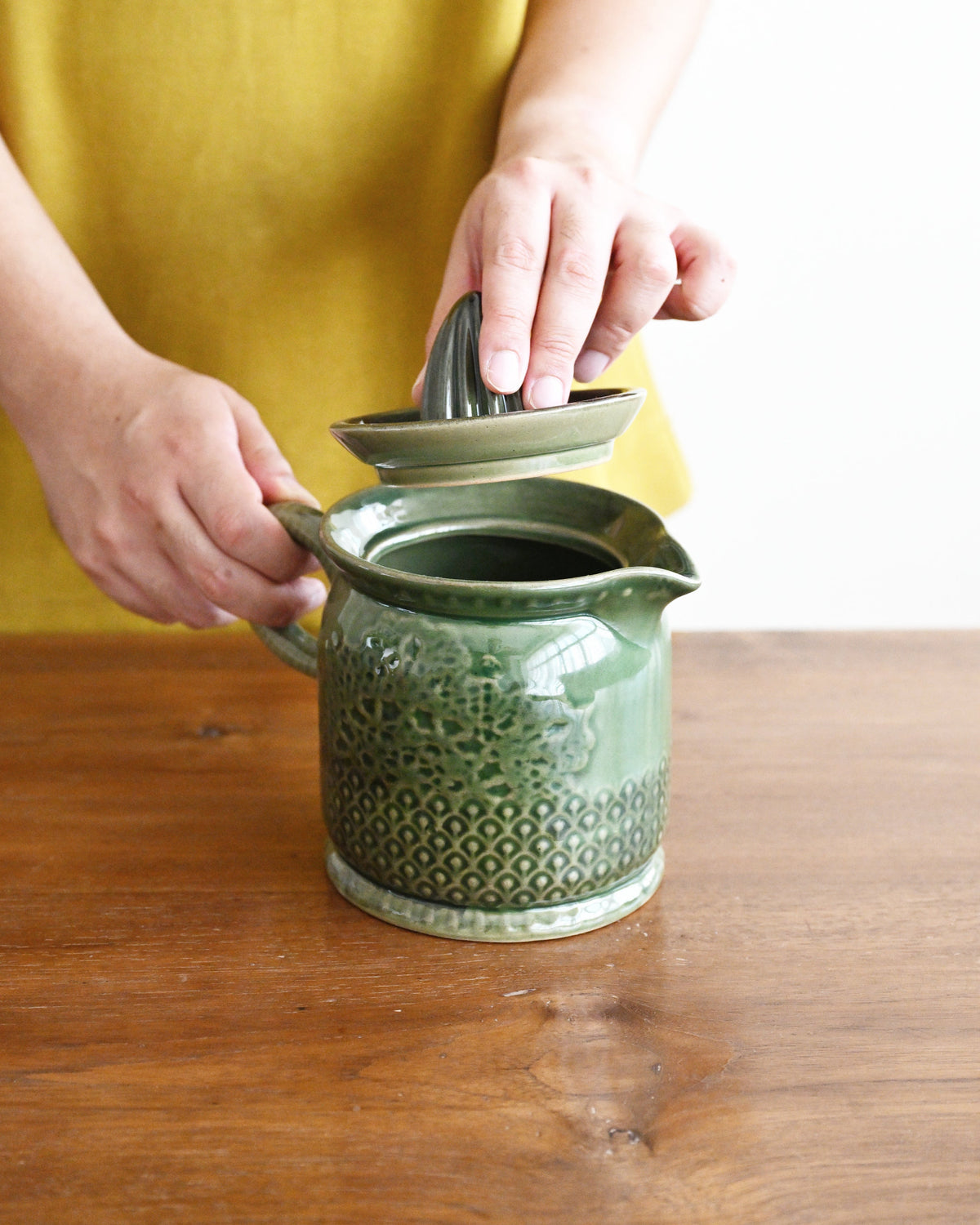 A person in a mustard yellow top holds the lid of a textured green ceramic pitcher on a wooden table.