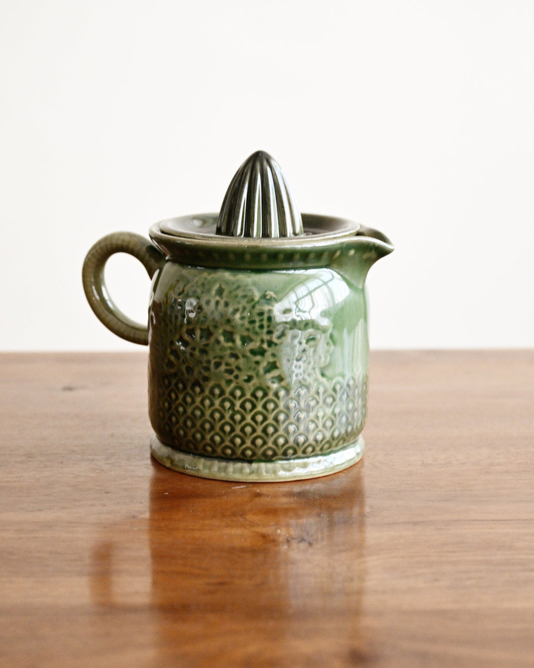 A small green ceramic citrus juicer with a textured pattern and a conical reamer on top, sitting on a wooden table against a plain white background.