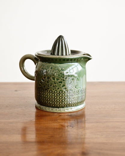 A small green ceramic citrus juicer with a textured pattern and a conical reamer on top, sitting on a wooden table against a plain white background.