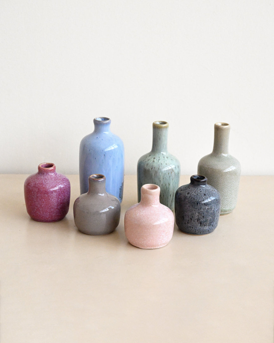 Seven small ceramic vases in assorted matte and textured glazes, including pink, blue, gray, green, and purple, arranged on a light beige surface against a neutral cream background.