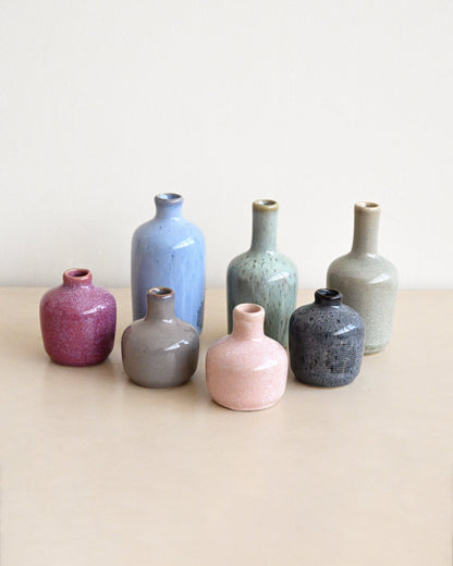 Seven small ceramic vases in assorted matte and textured glazes, including pink, blue, gray, green, and purple, arranged on a light beige surface against a neutral cream background.
