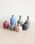 Seven small ceramic vases in assorted matte and textured glazes, including pink, blue, gray, green, and purple, arranged on a light beige surface against a neutral cream background.