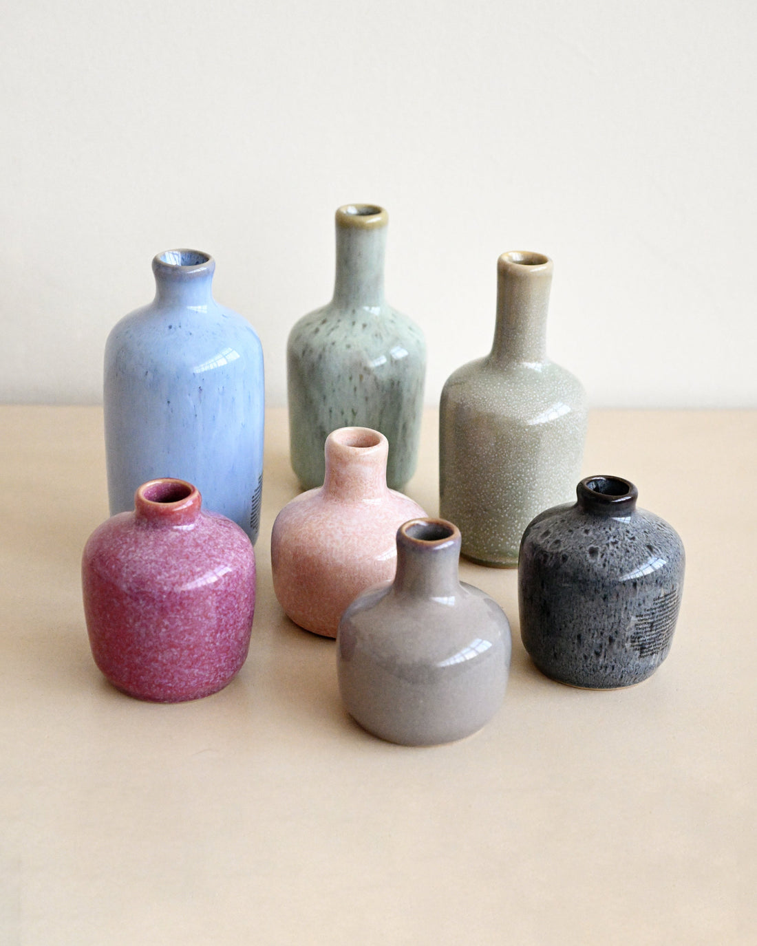 Six small ceramic vases in assorted matte and speckled glazes, including light blue, sage green, beige, magenta, pink, gray, and charcoal, arranged on a beige surface against a light neutral background.