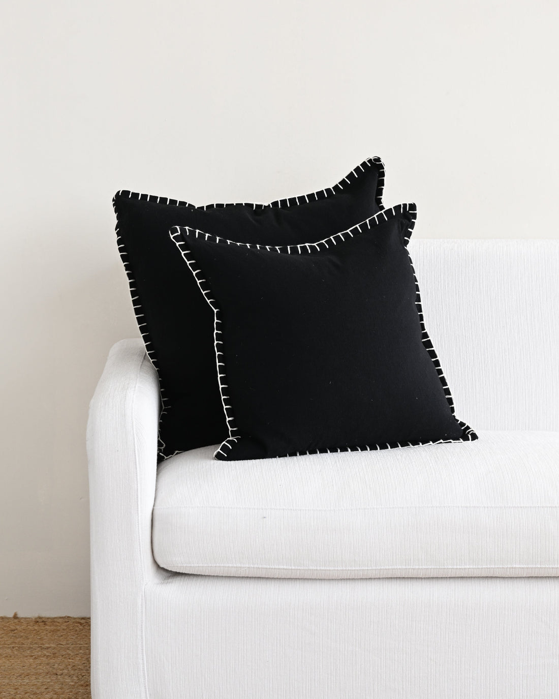 Two black pillows with white trim on a white couch against a light gray wall.