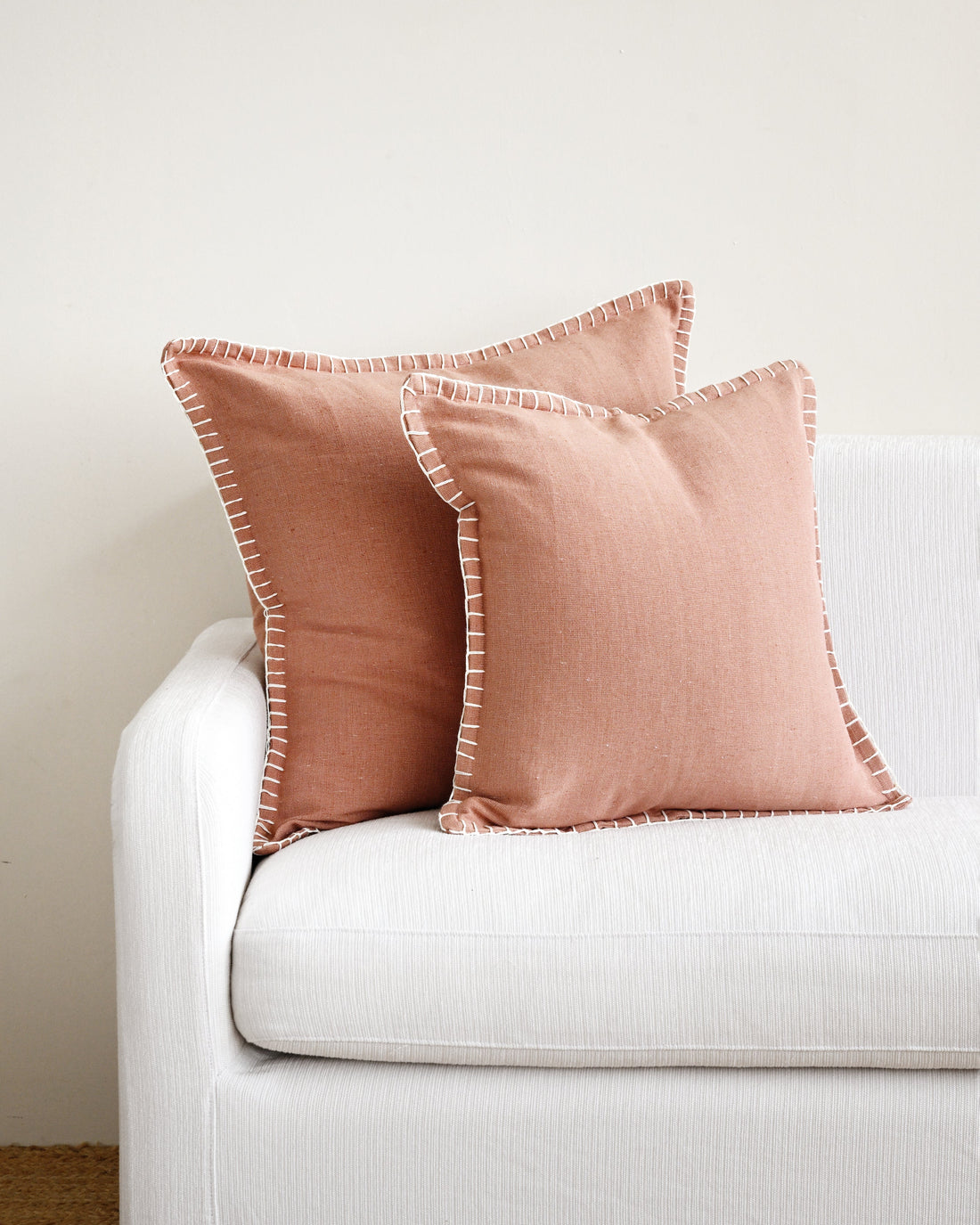 Two pink pillows with white trim on a white couch against a beige wall.