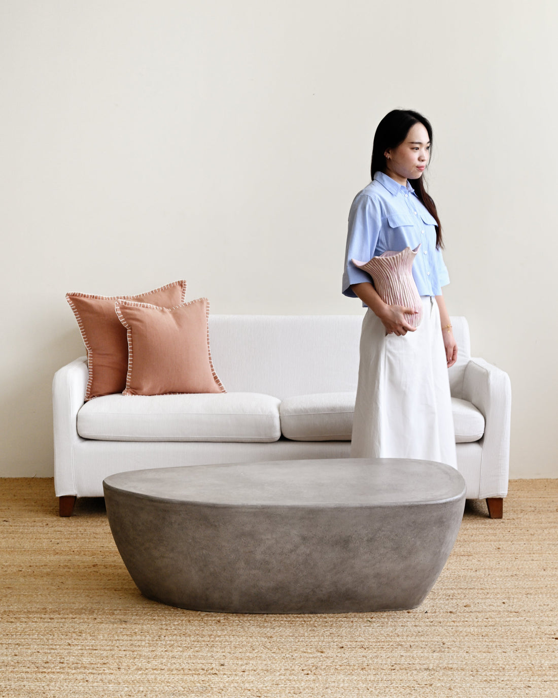Woman standing in a living room with a white sofa, pink pillows, and a concrete coffee table.