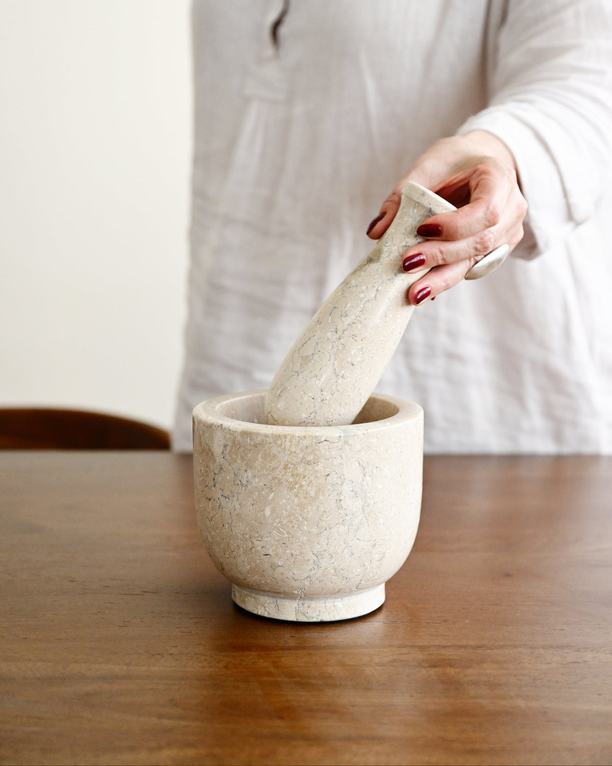 A person in a white linen top holds a matching stone mortar and pestle on a wooden table. The mortar and pestle have a speckled, textured off-white stone finish.