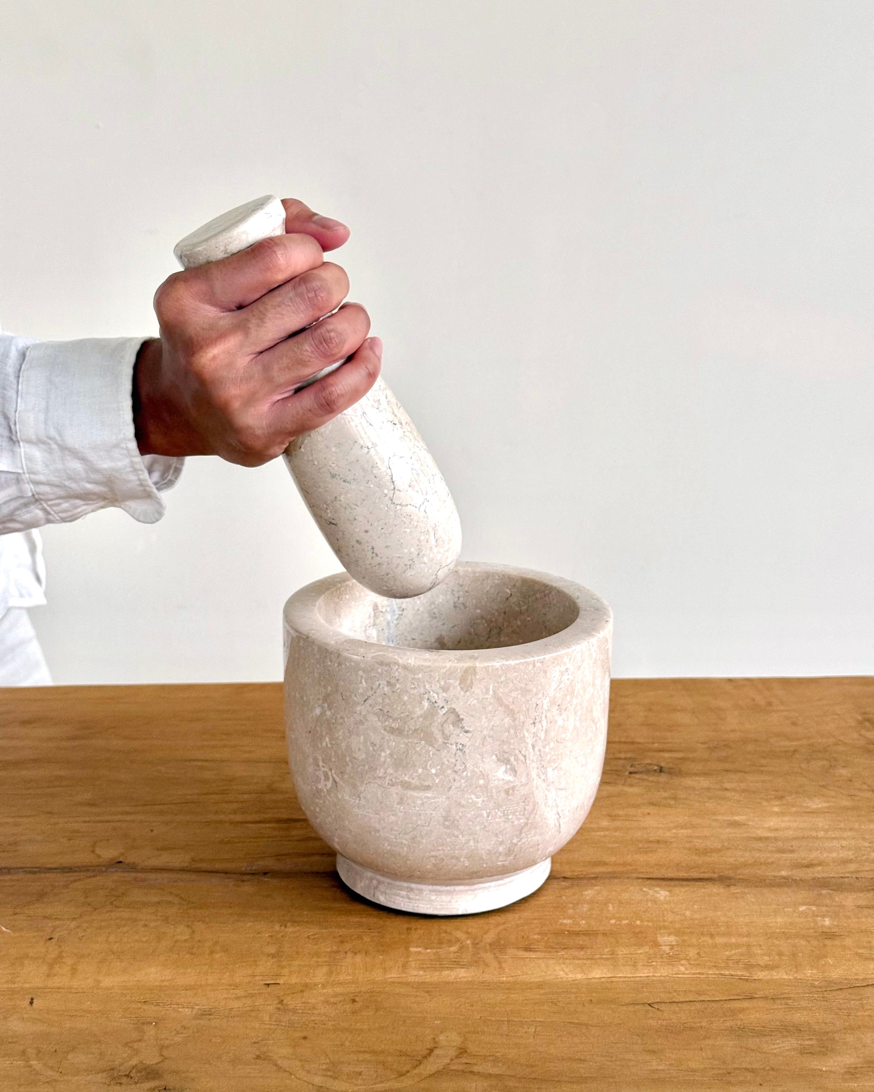 A hand holding a white stone pestle above a matching white stone mortar on a wooden table against a plain light background.