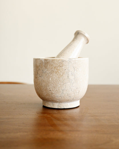 A light beige stone mortar and pestle set on a polished wooden table against a neutral cream background.