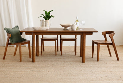 A mid-century modern wooden dining table set with four matching wooden chairs, styled with ceramic bowls, a vase of green leaves, and a wine bottle on a light beige woven rug against a white wall.