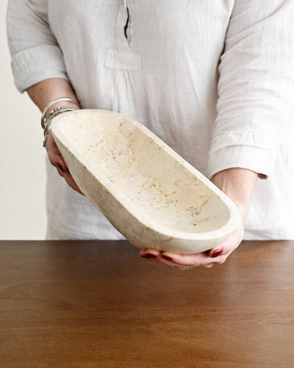 A person in a white linen shirt holds a carved oval stone bowl with a natural beige and cream marbled texture over a wooden table.