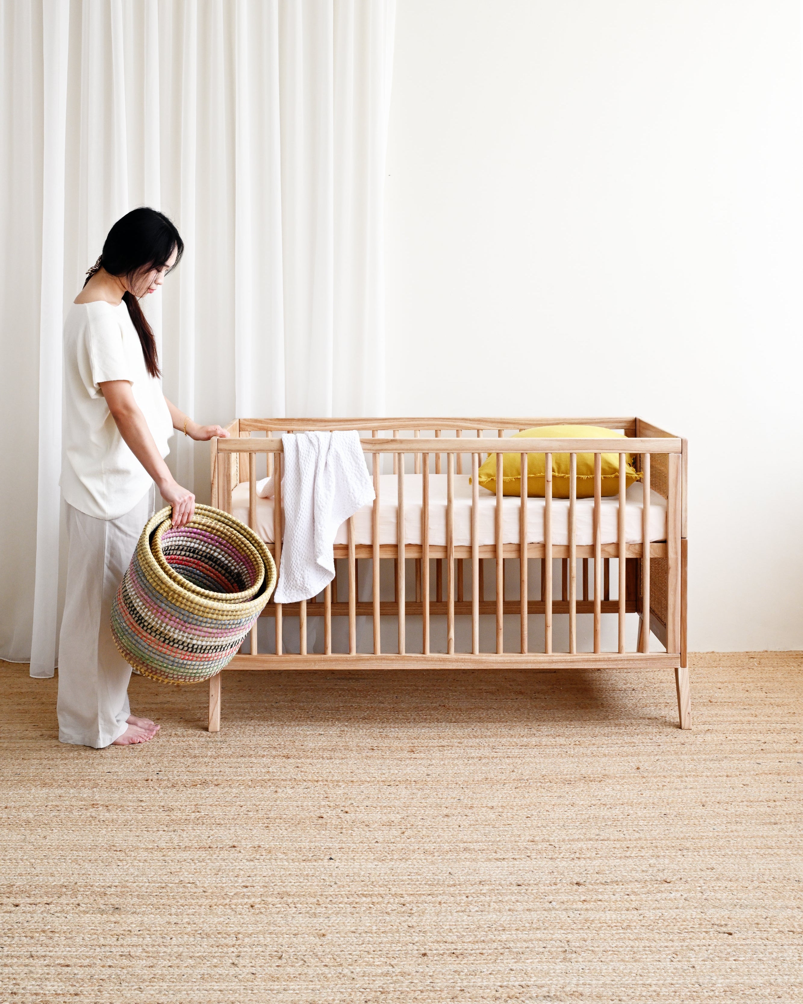 Woman holding a colorful basket next to a wooden crib in a room with white curtains.