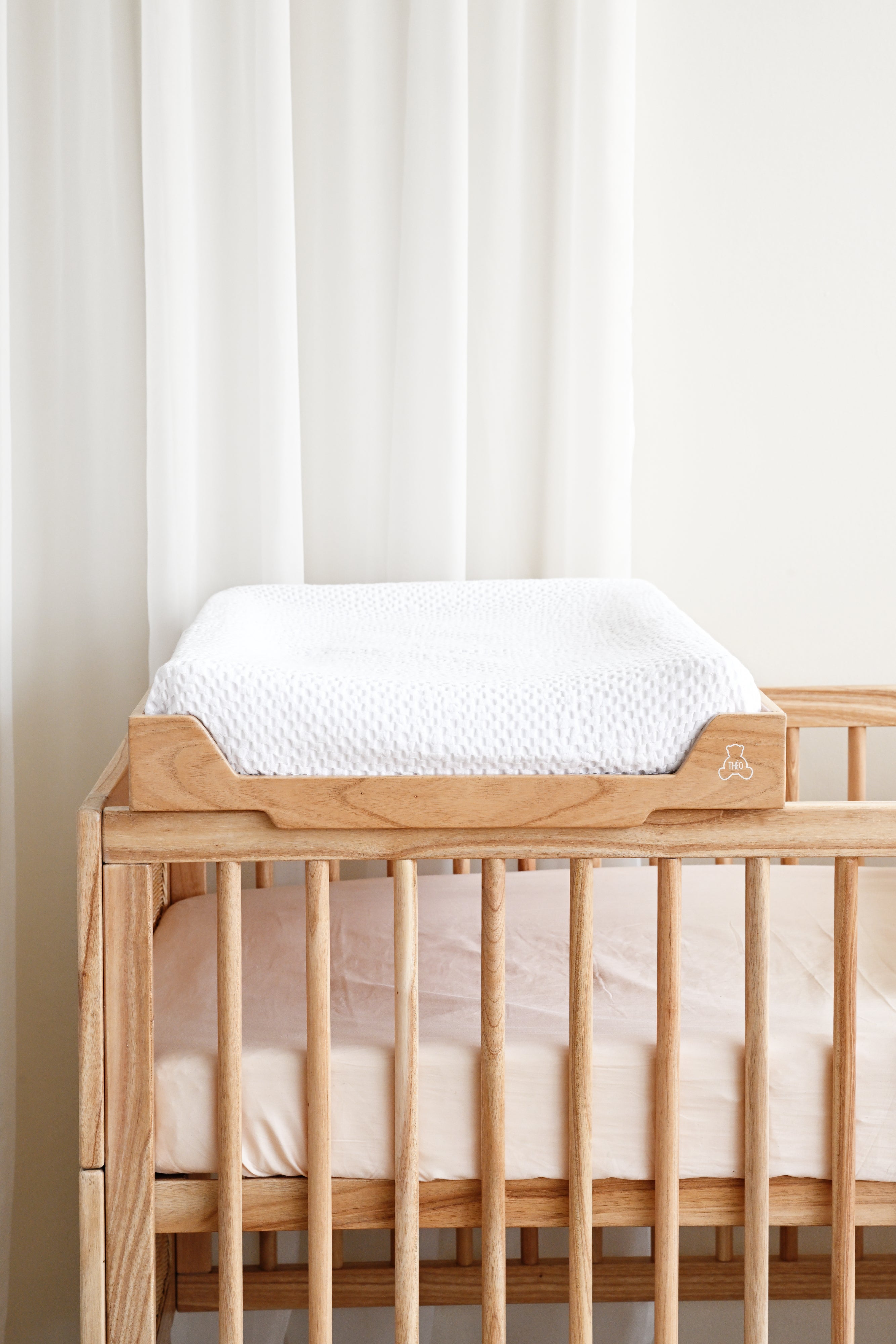 Wooden crib with a white mattress and fitted sheet against a white curtain background