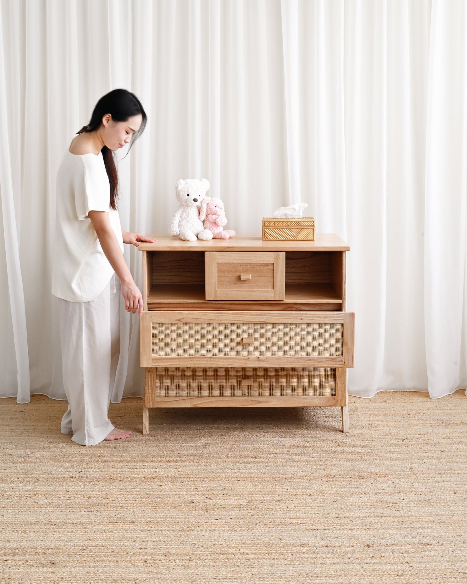 Woman standing next to a wooden dresser with a white teddy bear on top in a room with white curtains and beige carpet.