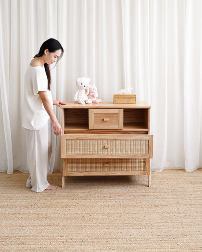 Woman standing next to a wooden dresser with a white teddy bear on top in a room with white curtains and beige carpet.