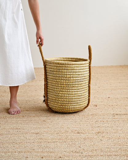 A woman in a white dress stands barefoot on a woven jute floor next to a tall, round woven basket with rope handles, set against a plain off-white wall.