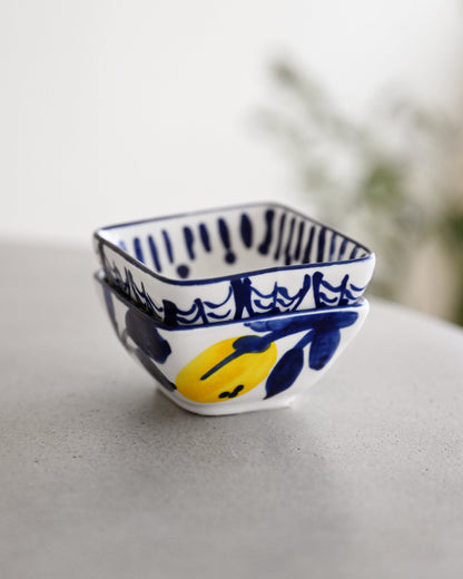 A small square white ceramic bowl with blue hand-painted patterns and a yellow lemon illustration, resting on a light gray surface with a soft-focus background.