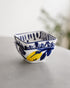 A small square white ceramic bowl with blue hand-painted patterns and a yellow lemon illustration, resting on a light gray surface with a soft-focus background.