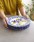 A person in a mustard yellow shirt holds a stack of ceramic plates with a white background, blue floral and geometric border patterns, and small yellow lemon illustrations.
