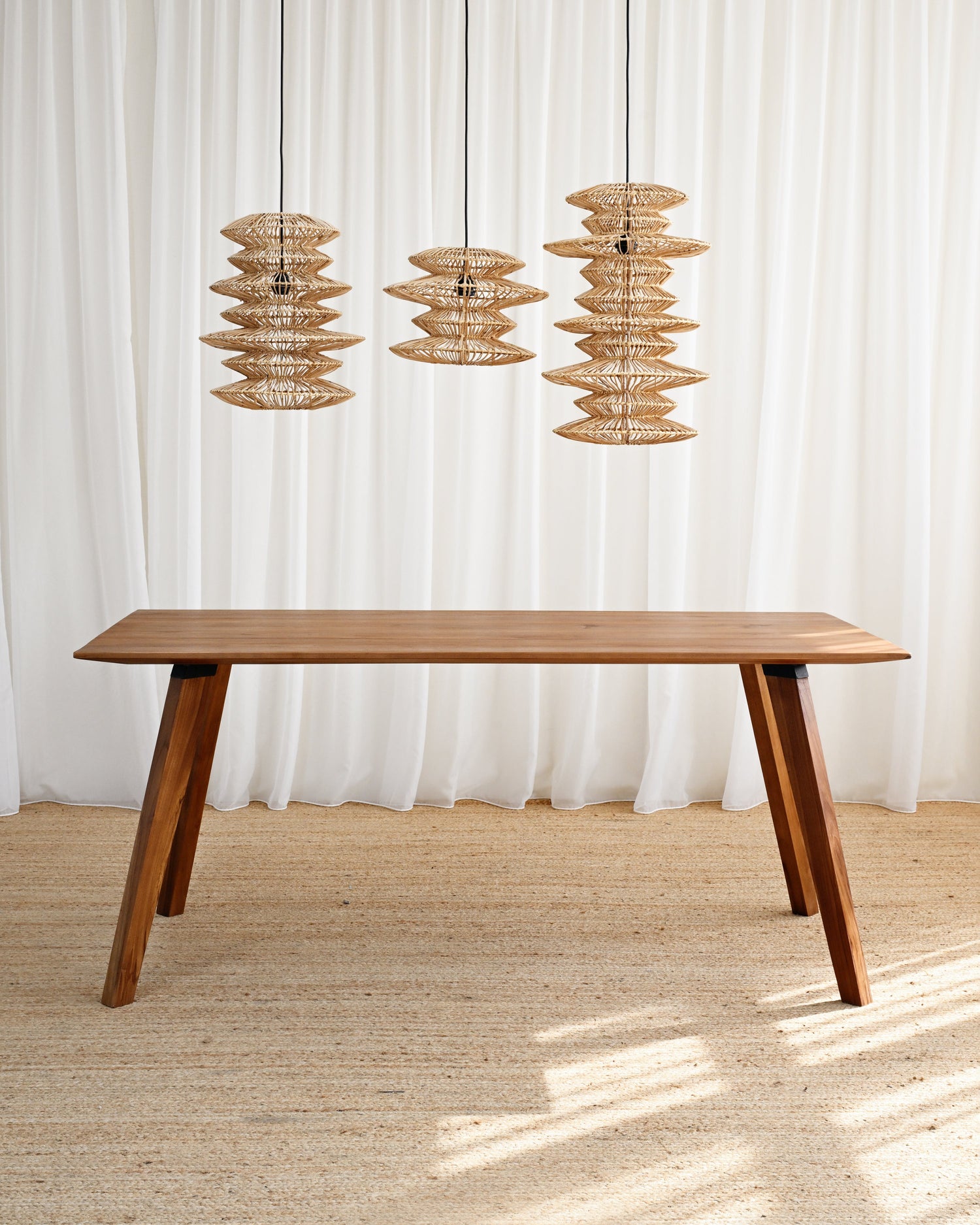 Wooden dining table with three hanging wicker lamps against a white curtain background