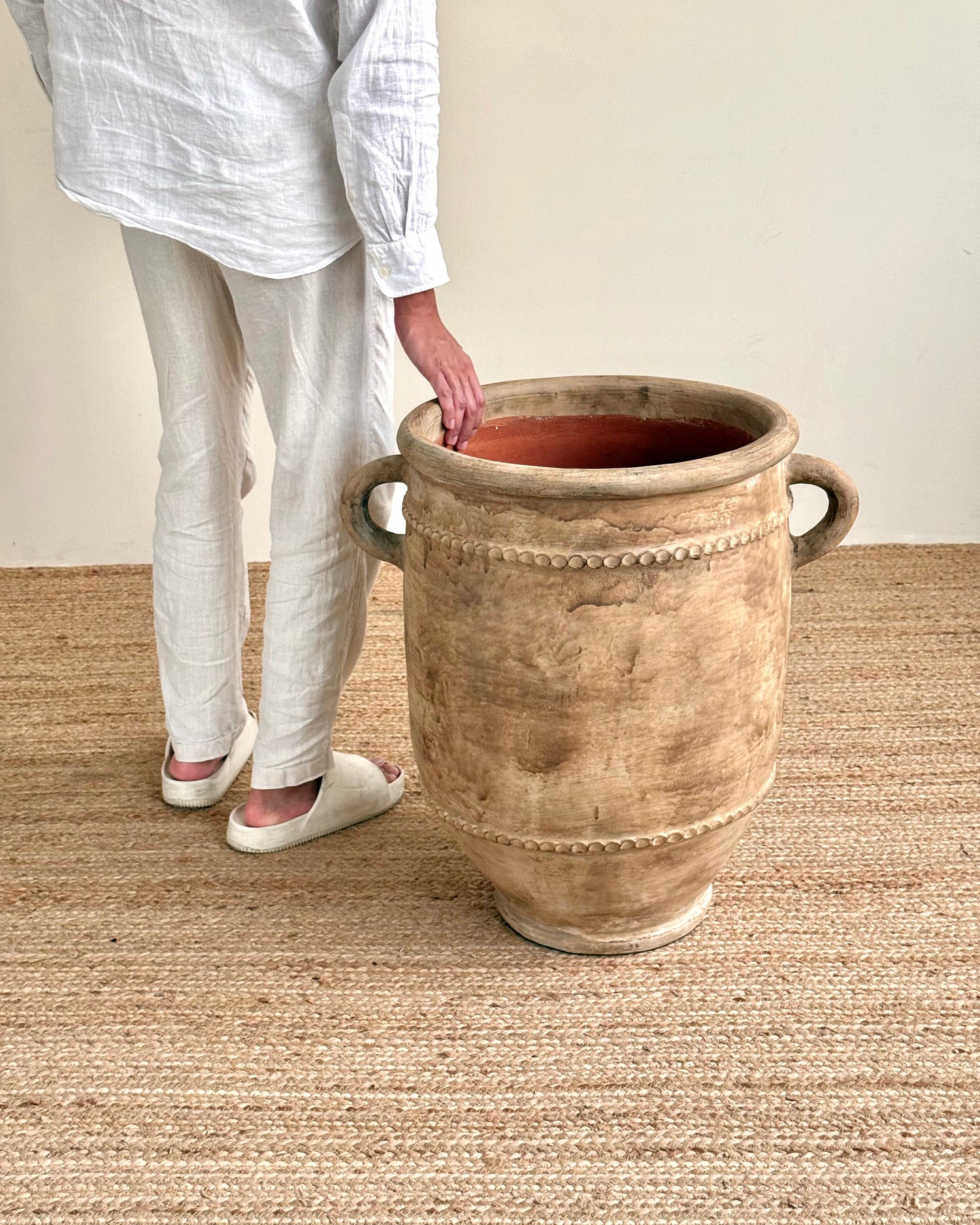 A person wearing a white linen shirt and pants stands beside a large rustic beige ceramic planter pot with side handles on a woven jute rug.