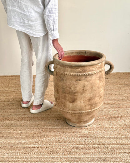 A person wearing a white linen shirt and pants stands beside a large rustic beige ceramic planter pot with side handles on a woven jute rug.