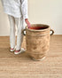 A person wearing a white linen shirt and pants stands beside a large rustic beige ceramic planter pot with side handles on a woven jute rug.