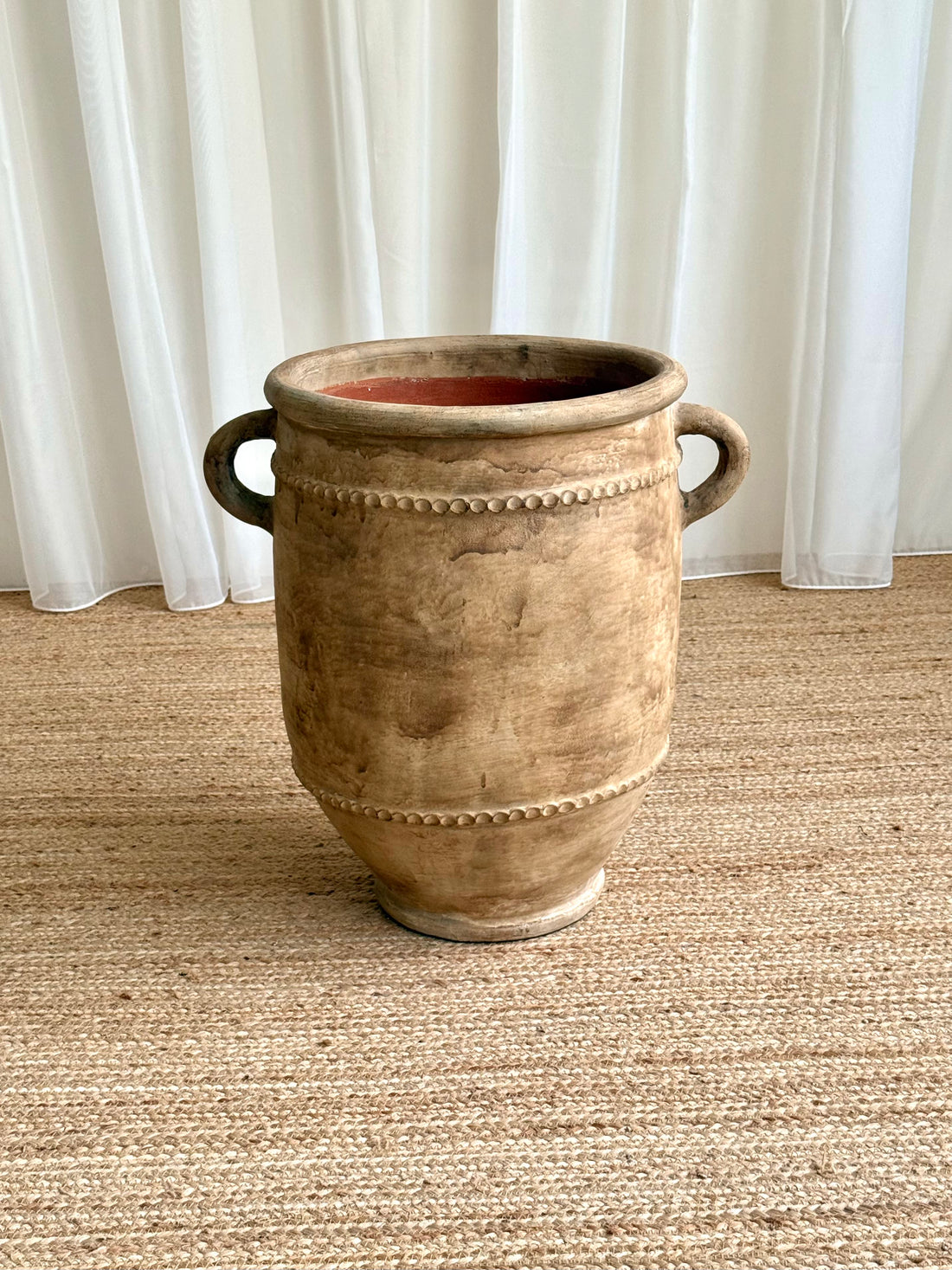 A large rustic ceramic planter pot with two side handles and decorative beaded bands, sitting on a woven jute rug in front of white curtains.