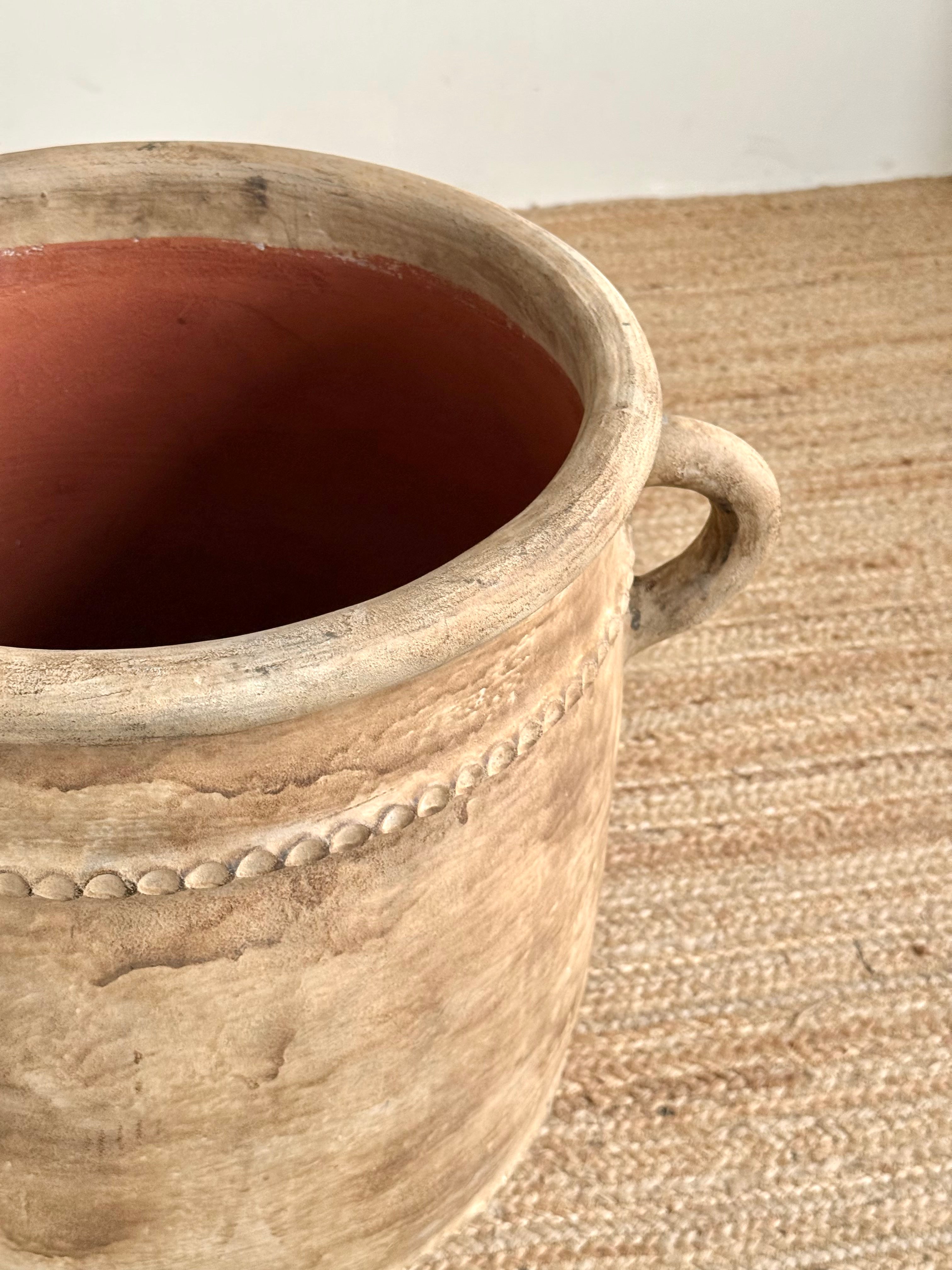 A rustic ceramic planter pot with a textured beige exterior, a small loop handle on the right side, and a reddish-brown interior, shown on a woven jute rug.
