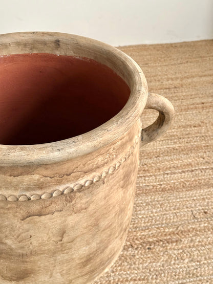 A rustic ceramic planter pot with a textured beige exterior, a small loop handle on the right side, and a reddish-brown interior, shown on a woven jute rug.