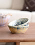 A small handmade ceramic bowl with a blue and yellow speckled glaze, stacked on a wooden table in a softly blurred living room setting.