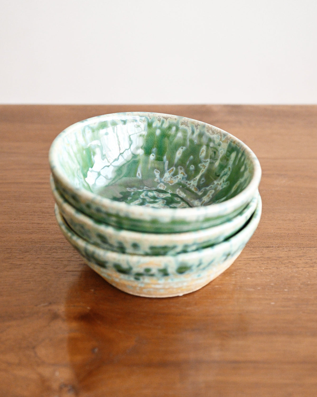 A stack of four handmade ceramic bowls with a green and white marbled glaze, displayed on a wooden tabletop against a neutral off-white background.