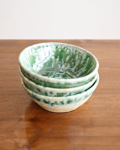 A stack of four handmade ceramic bowls with a green and white marbled glaze, displayed on a wooden tabletop against a neutral off-white background.