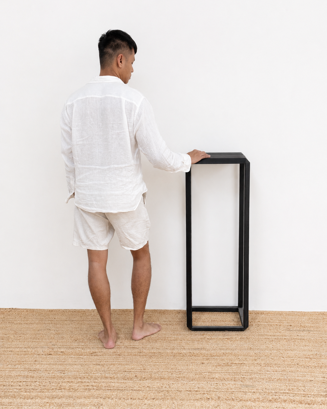 Man in white outfit standing next to a black minimalist table against a white wall.