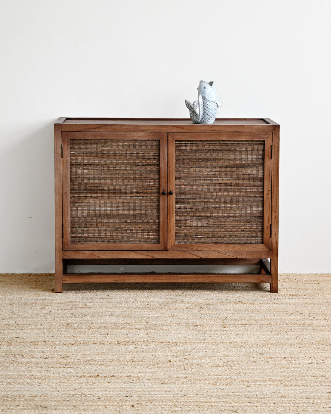 A mid-century modern wooden credenza or sideboard cabinet with two woven rattan doors, shown on a textured jute rug against a plain white wall. A small gray ceramic bird figurine sits on top.